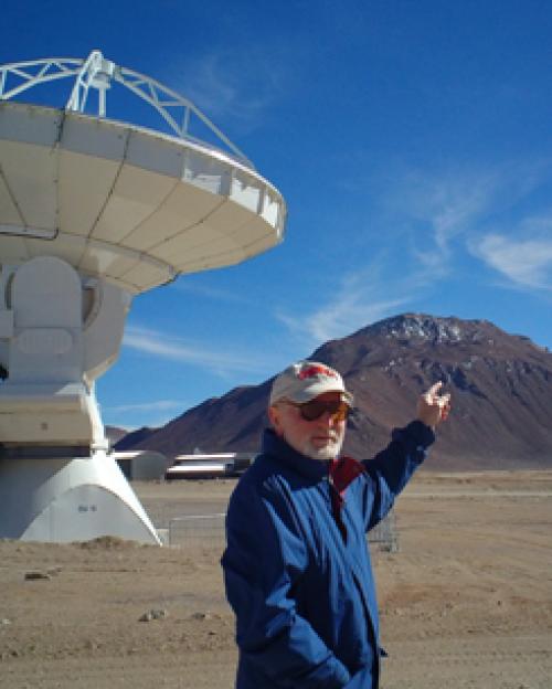 Riccardo Giovanelli in blue jacket, baseball cap, sunglasses and white beard, points to the top of a mountain in the desert. A telescope is seen in the background.