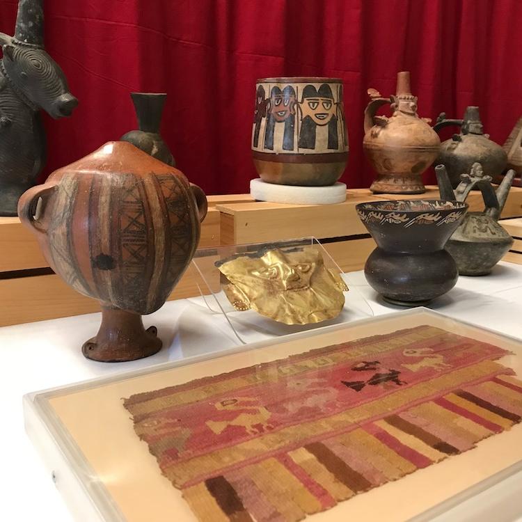 A display case showing a ceramic head-shaped object standing on a base; a woven cloth showing animal shapes; and a gold mask