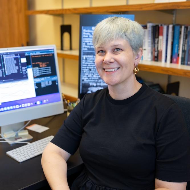 		Person sitting at a desk with books in the background and graphs on a computer screen 
	