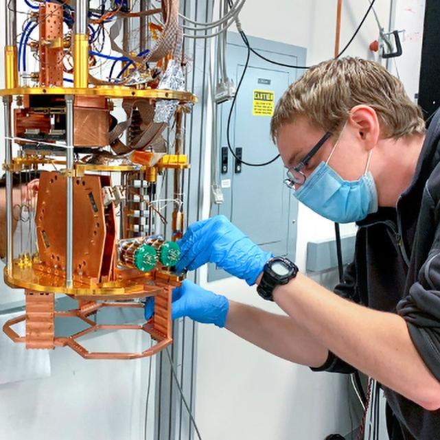 		Two people lean toward a complicated scientific instrument featuring gold and orange metal parts; it's about the size of a coffeemaker
	