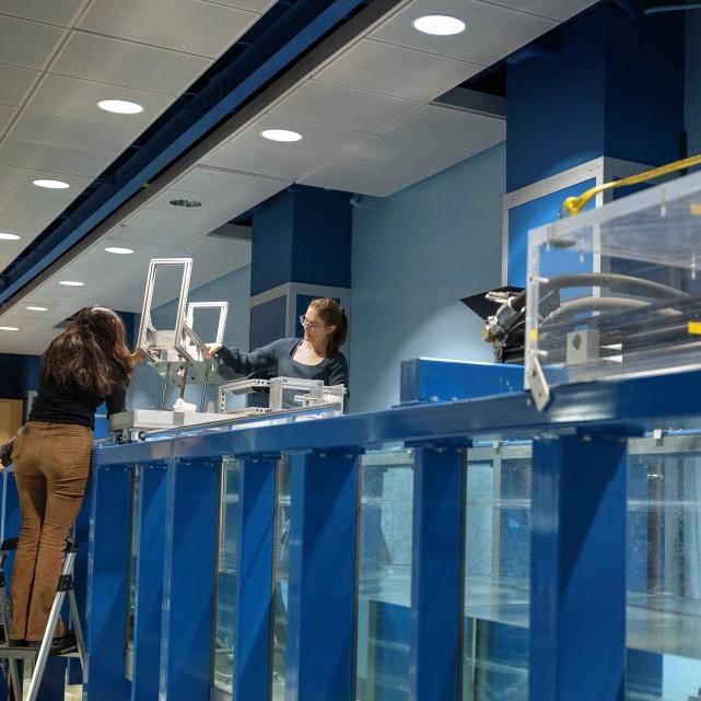Two people work on a very large piece of equipment that's blue and holds a lot of water