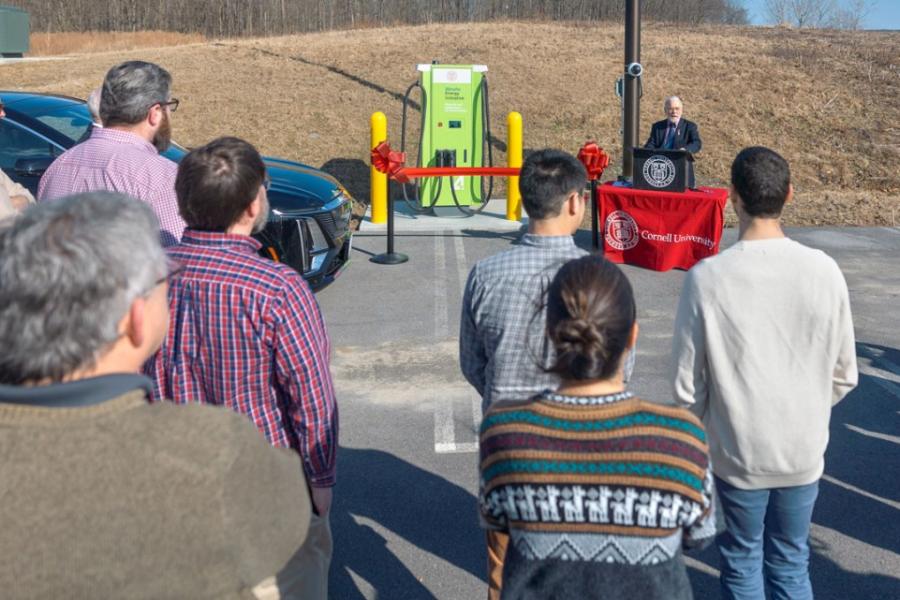 Héctor D. Abruña gives remarks Sevearl people stand in a parking lot, listening to a speaker who stands at a makeshift podium next to a large green machine