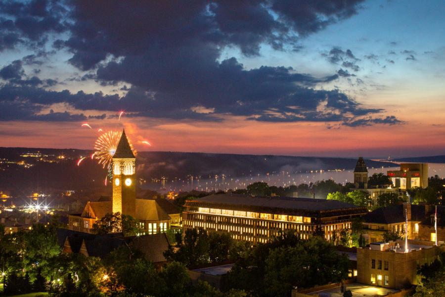 A dramatic night sky behind a college campus; a burst of fireworks explodes behind a clock tower with illuminated windows