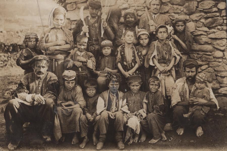 Historic photo of about 20 people, many of them children arranged in rows against a stone wall of a building