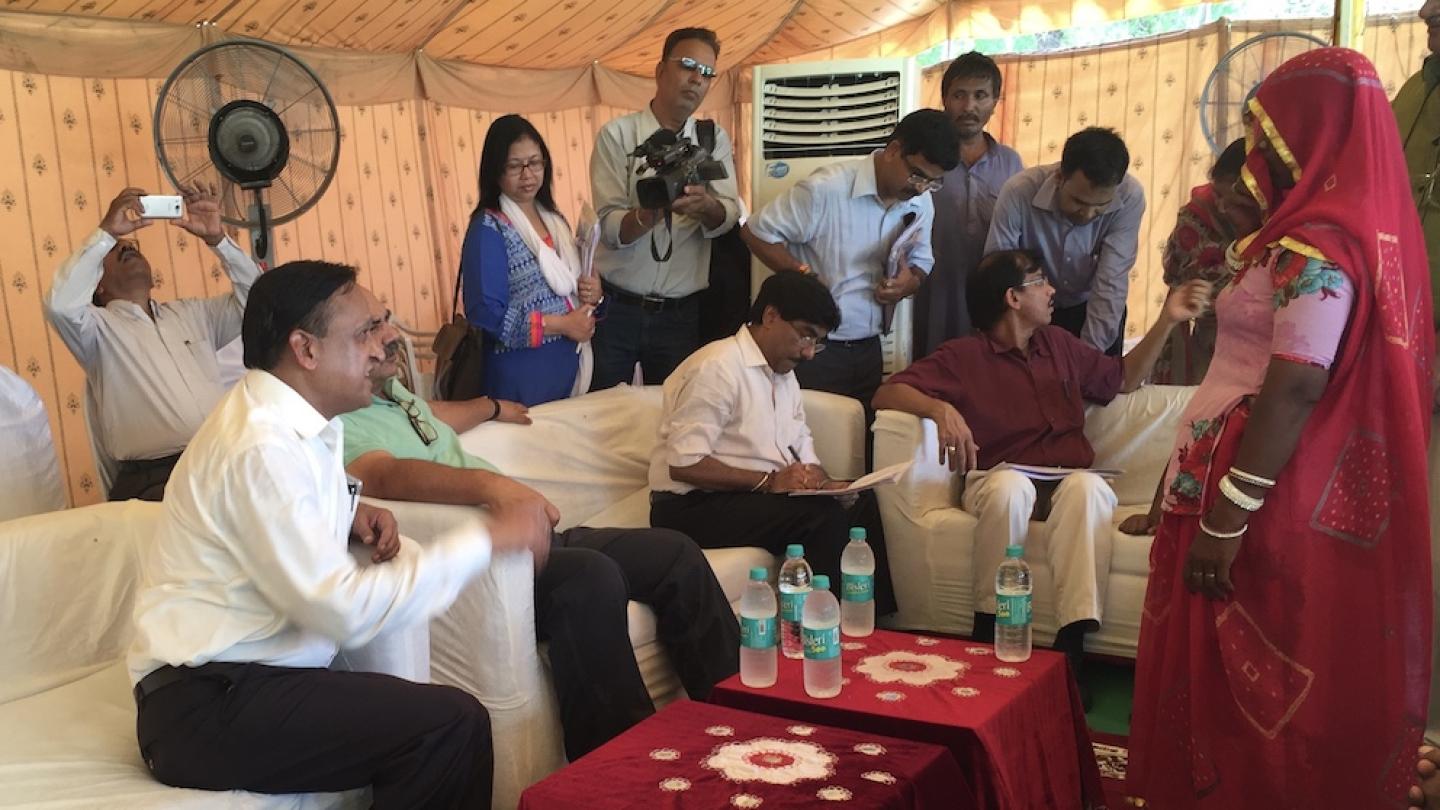 Among several people in a tent, a woman in red traditional clothing faces three officials in white shirts and black trousers, sitting on a couch