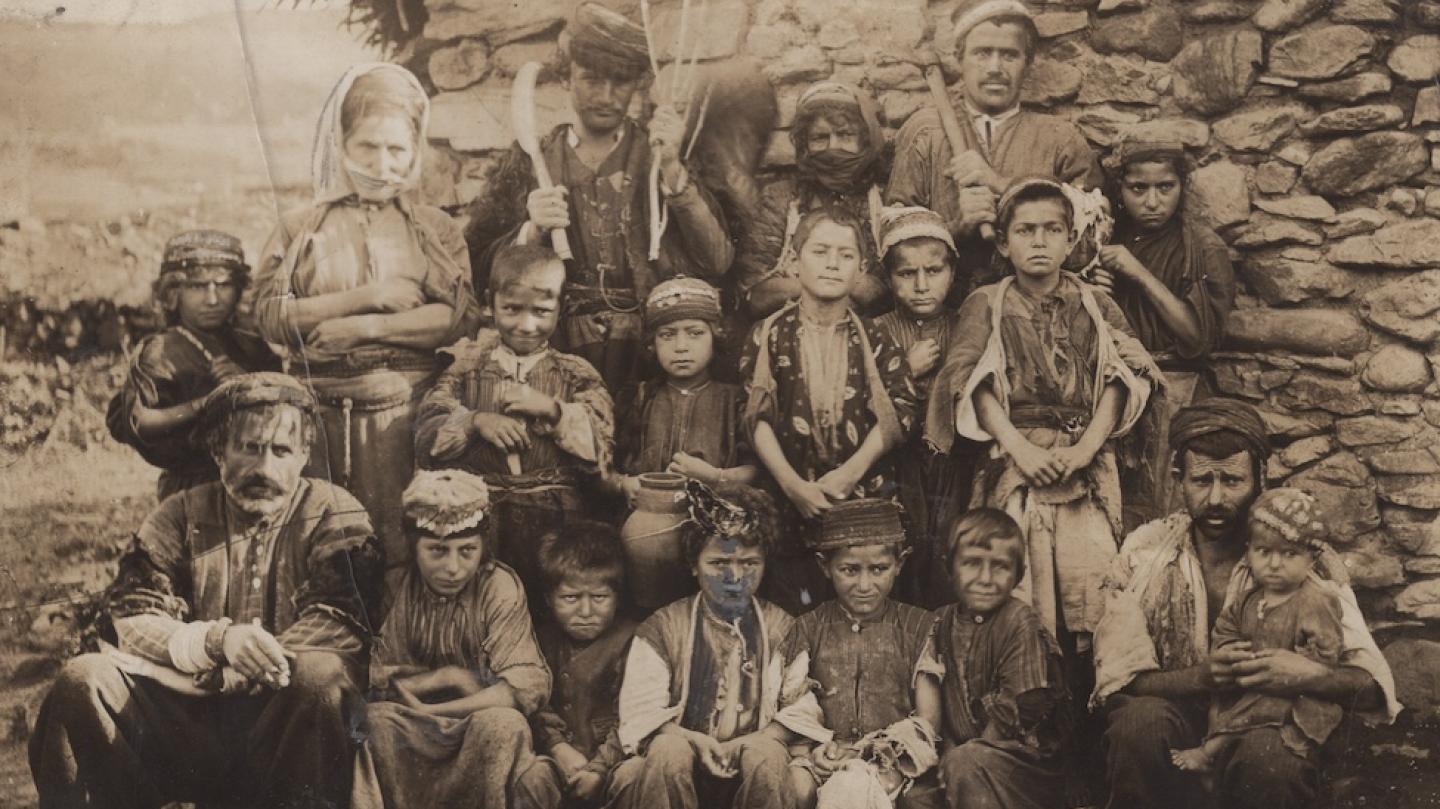 Historic photo of about 20 people, many of them children arranged in rows against a stone wall of a building